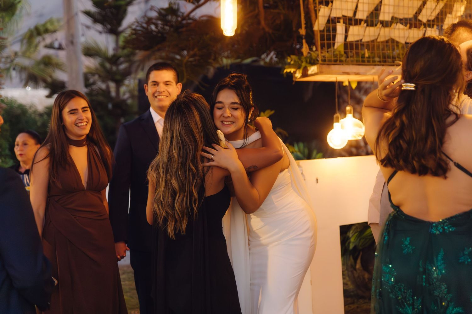 Bride in white wedding dress warmly hugging a guest during an outdoor evening wedding reception celebration.