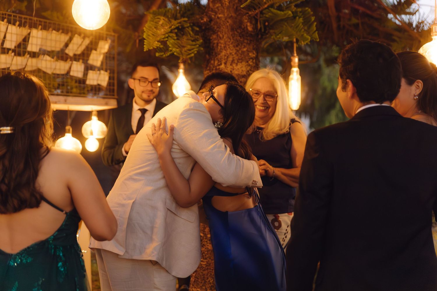 Two women share a warm hug at an outdoor evening wedding reception under warm string lights.