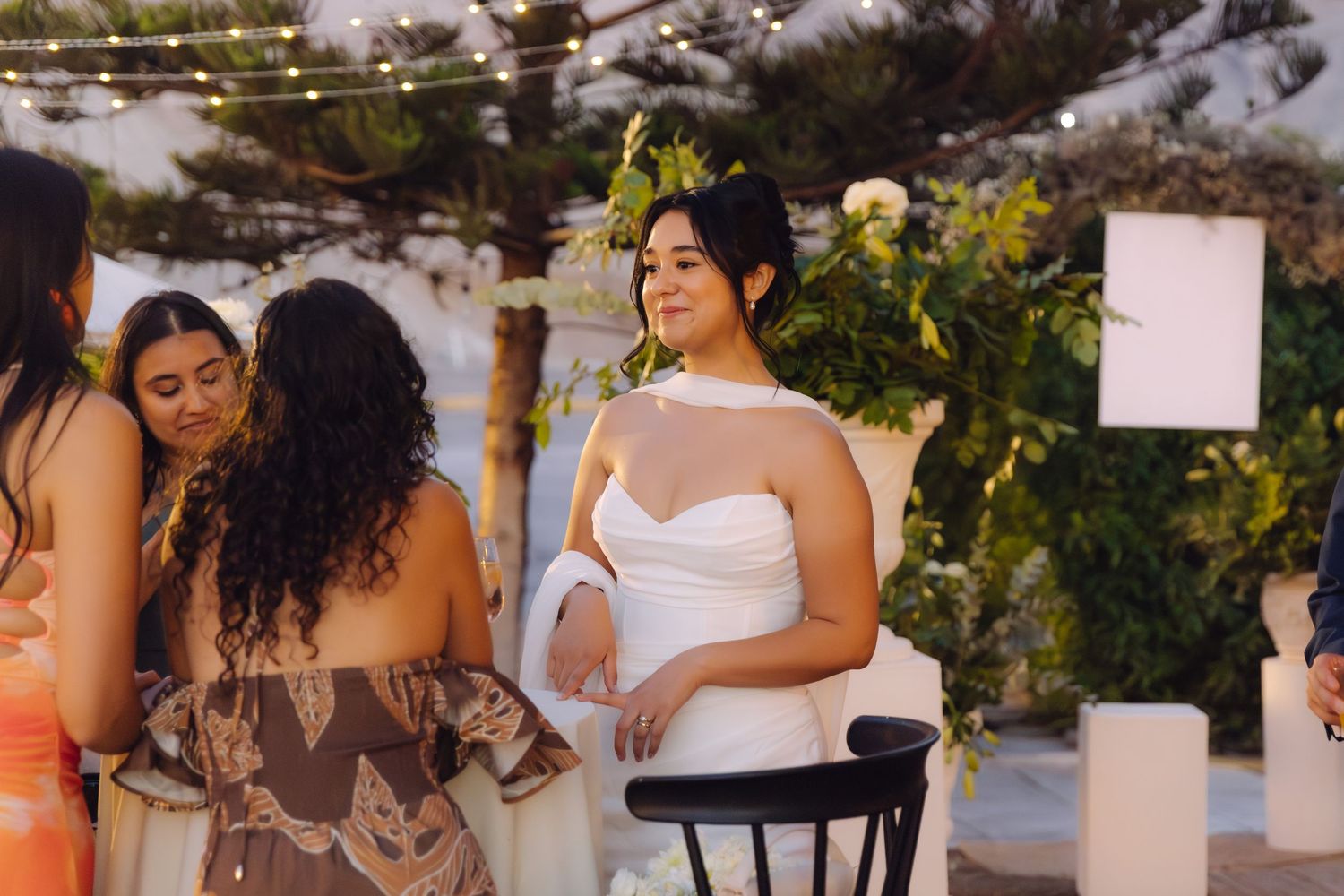 Elegant bride in white strapless gown sits gracefully at an outdoor wedding reception.