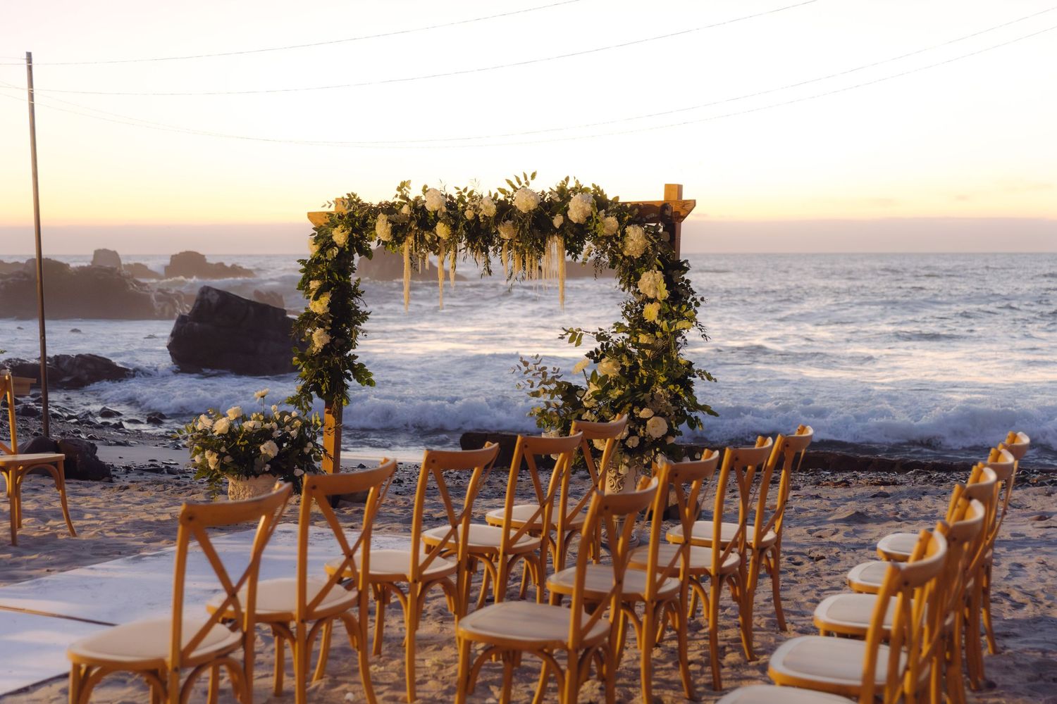 Romantic beach wedding ceremony setup with floral arch and wooden chairs at sunset by the ocean.