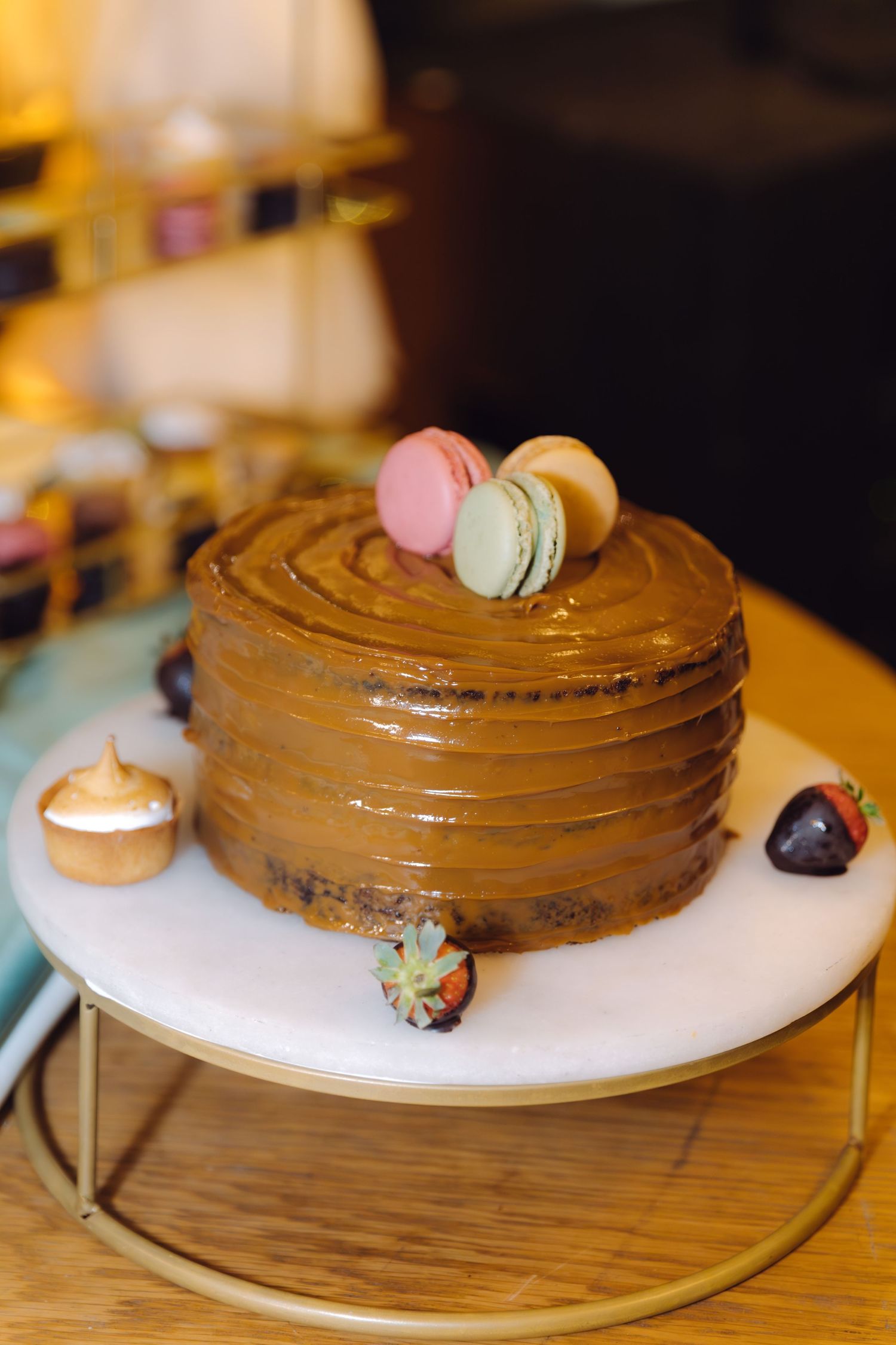 Elegant chocolate cake with macarons on top, decorated with chocolate-dipped strawberries on a marble cake stand.