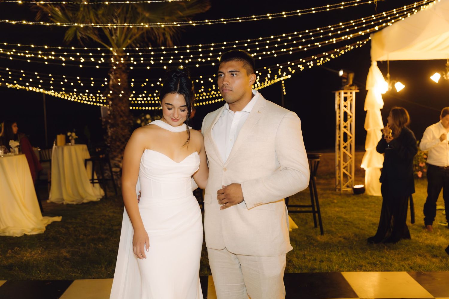 Bride in white strapless gown and groom in beige suit pose under fairy lights at outdoor wedding reception.
