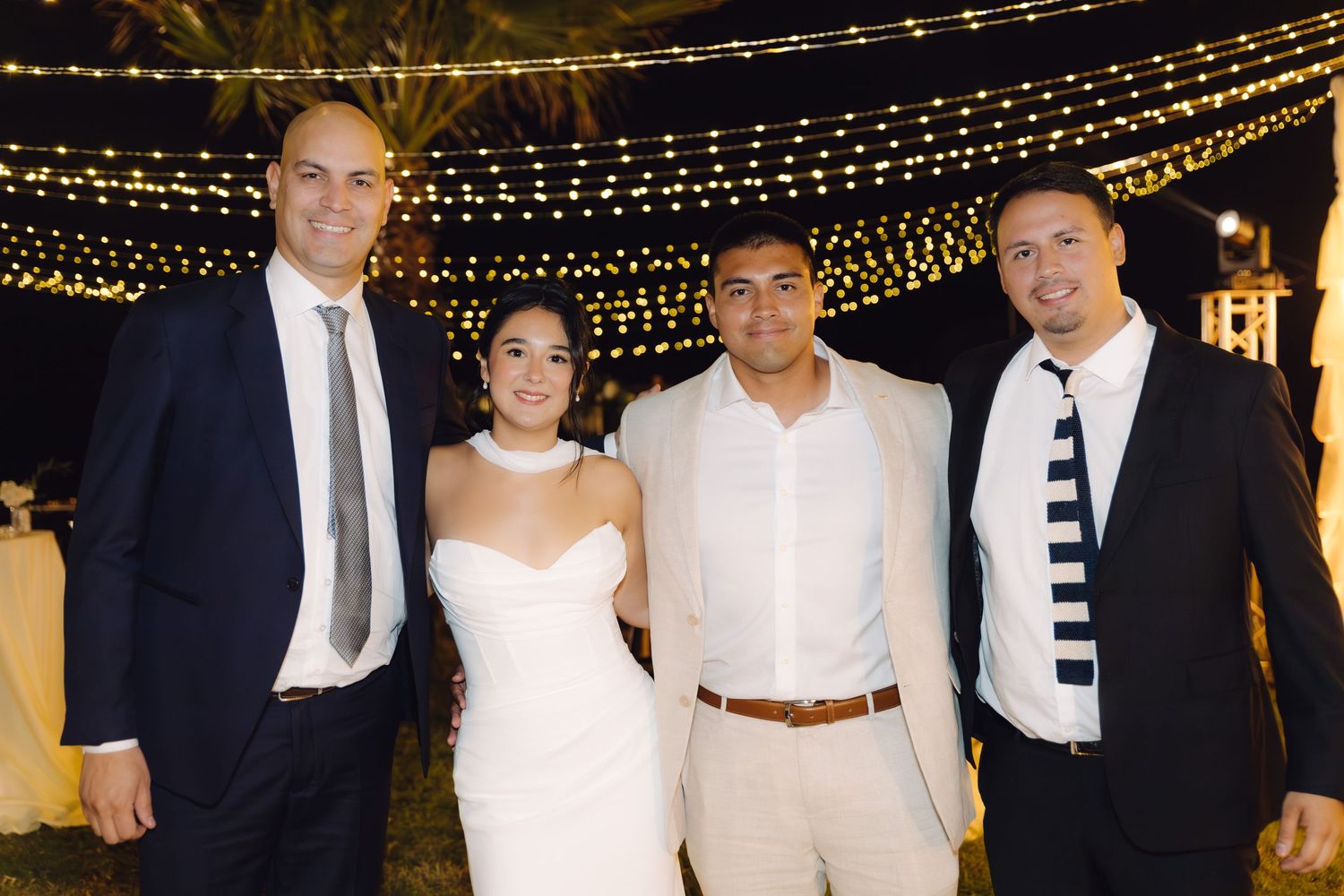 Bride and groom pose with two male wedding guests under twinkling fairy lights at elegant outdoor reception.