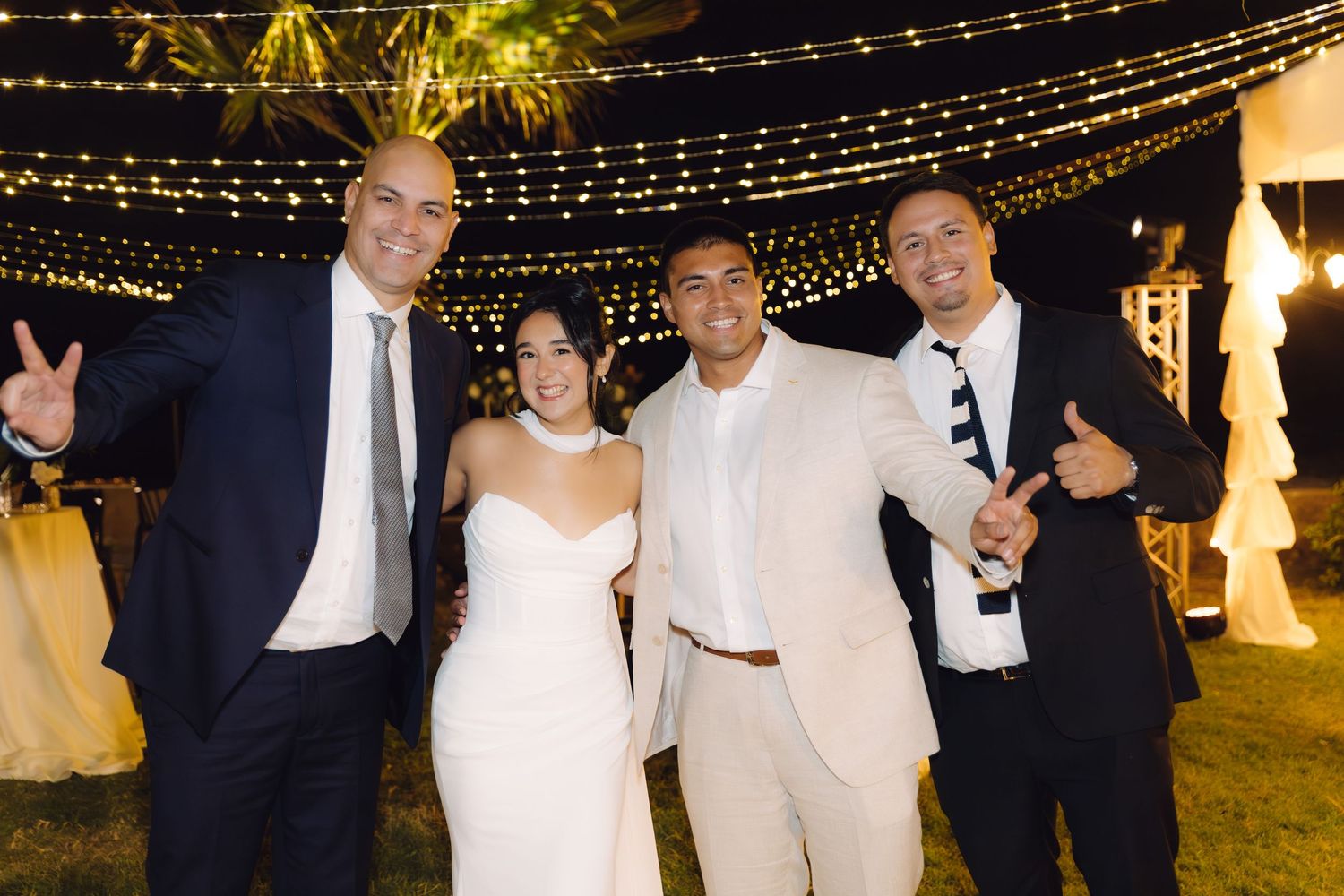 Smiling bride and groom celebrate with two male guests making fun poses under fairy lights at wedding.