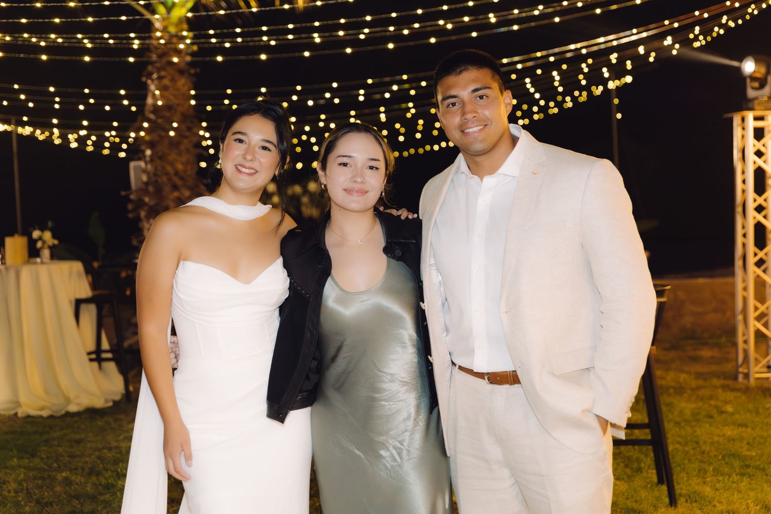 Bride in white dress and groom in beige suit pose with female guest in silver dress at night wedding.
