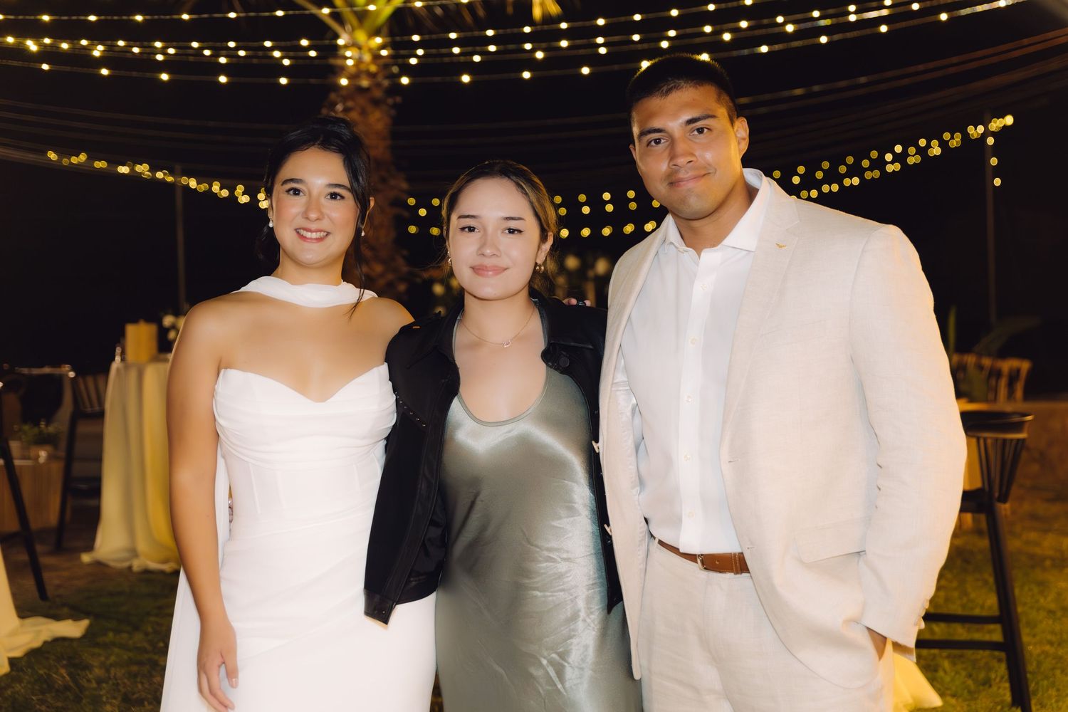 Bride, groom, and female guest in silver dress smile together under string lights at outdoor wedding.