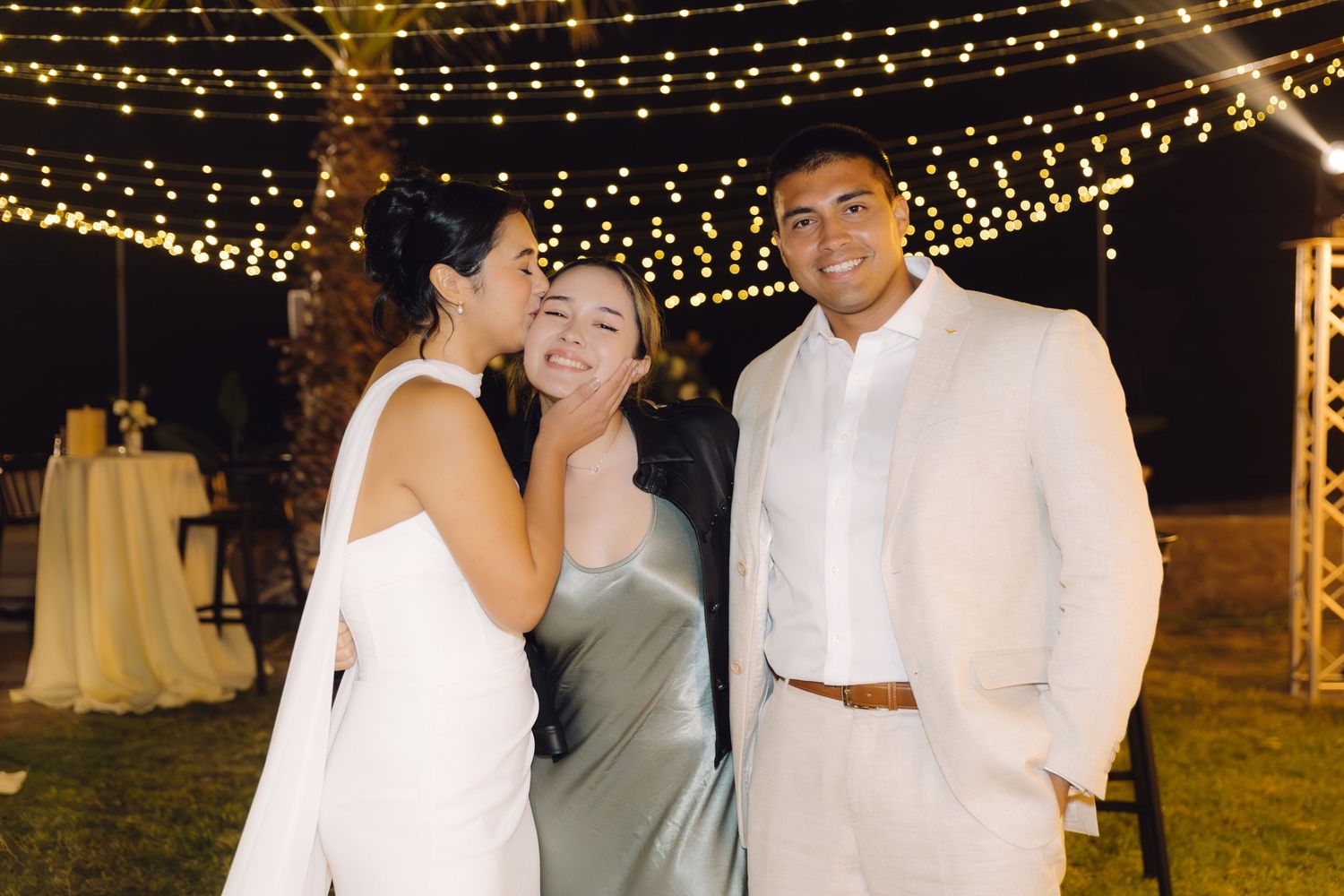 Bride laughing with bridesmaid in silver dress while groom smiles at romantic outdoor wedding reception.