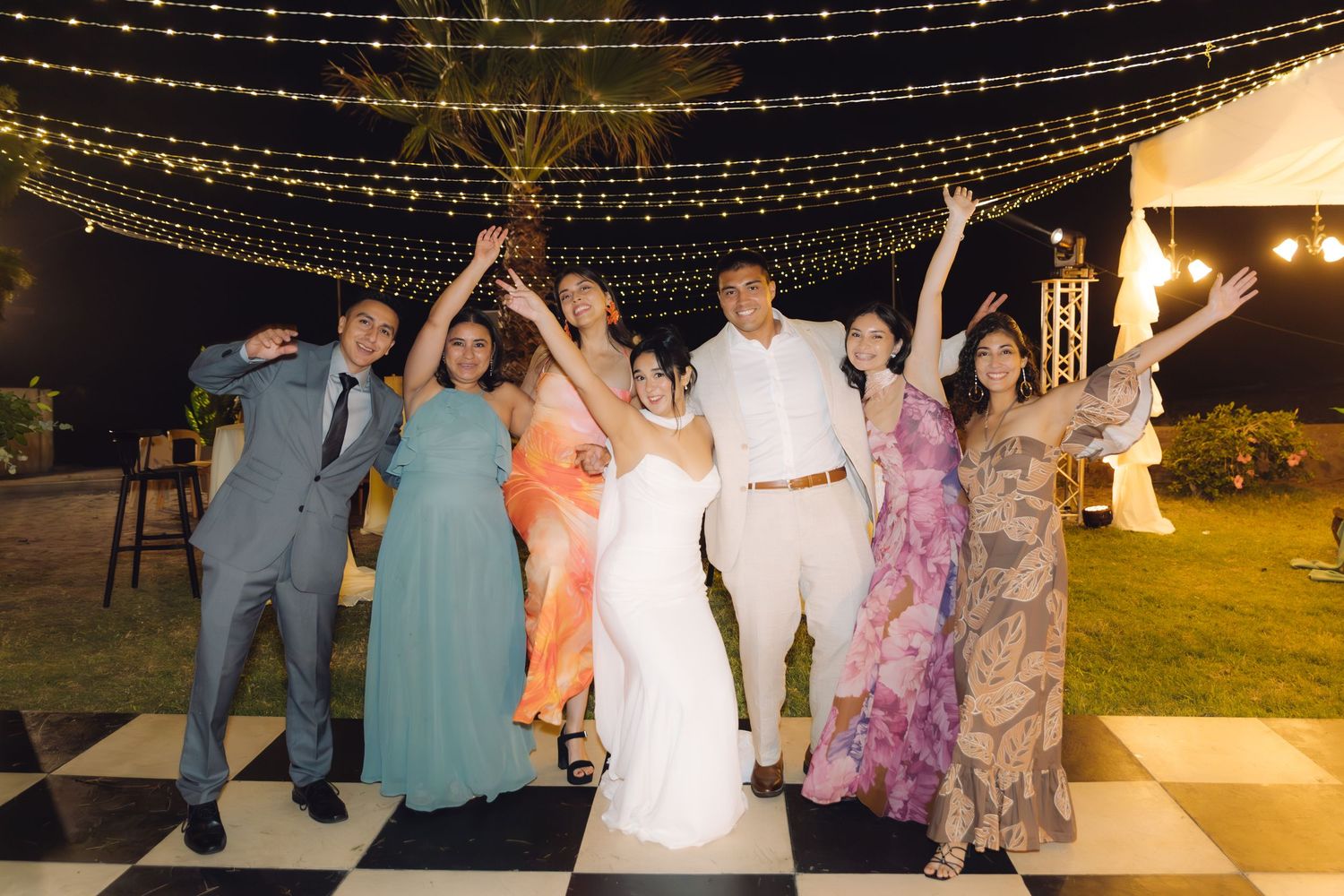 Wedding party celebrating joyfully on black and white dance floor under fairy lights at outdoor reception.