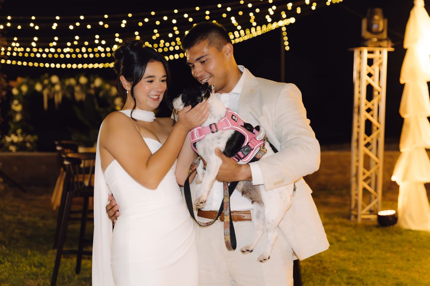 Bride and groom lovingly hold small dog in pink outfit under string lights at outdoor wedding reception.