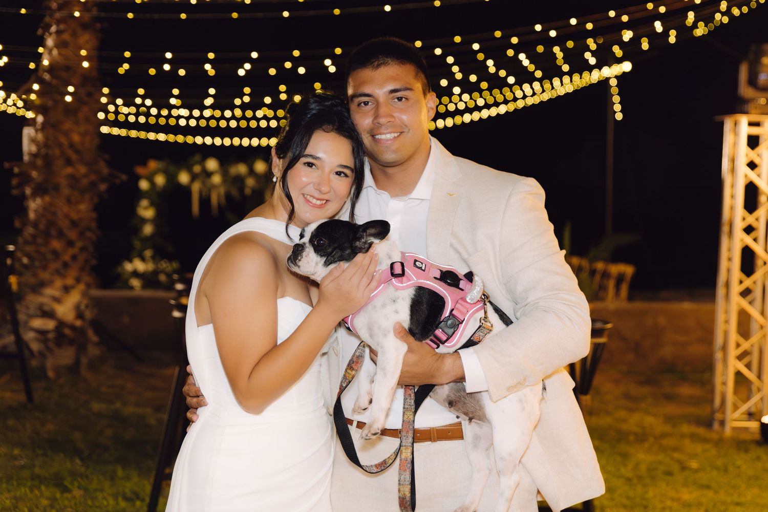Newlyweds smiling and holding small dog dressed in pink outfit under fairy lights at outdoor wedding.