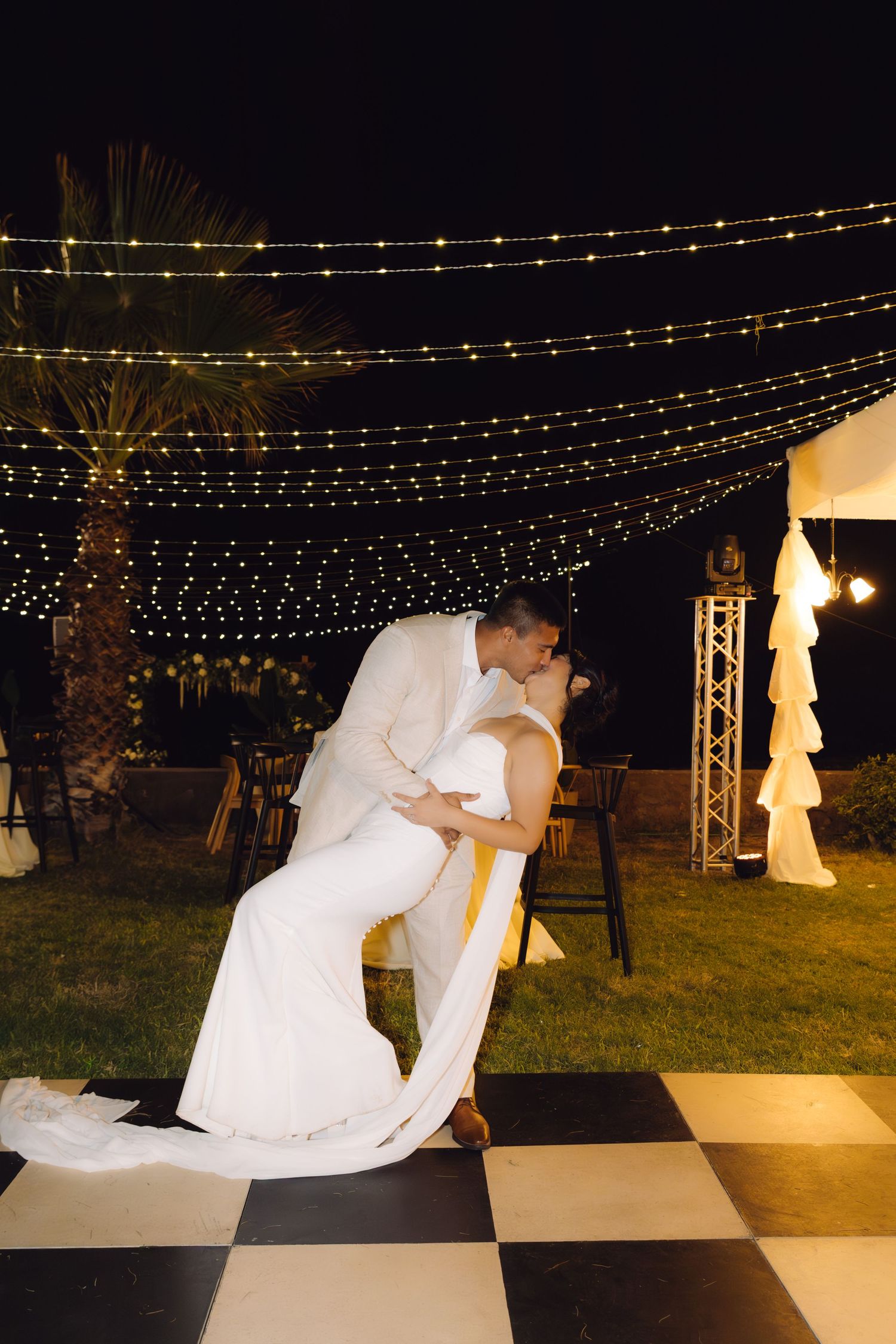 Bride and groom share a romantic dip kiss on a black and white checkered dance floor under string lights at night.