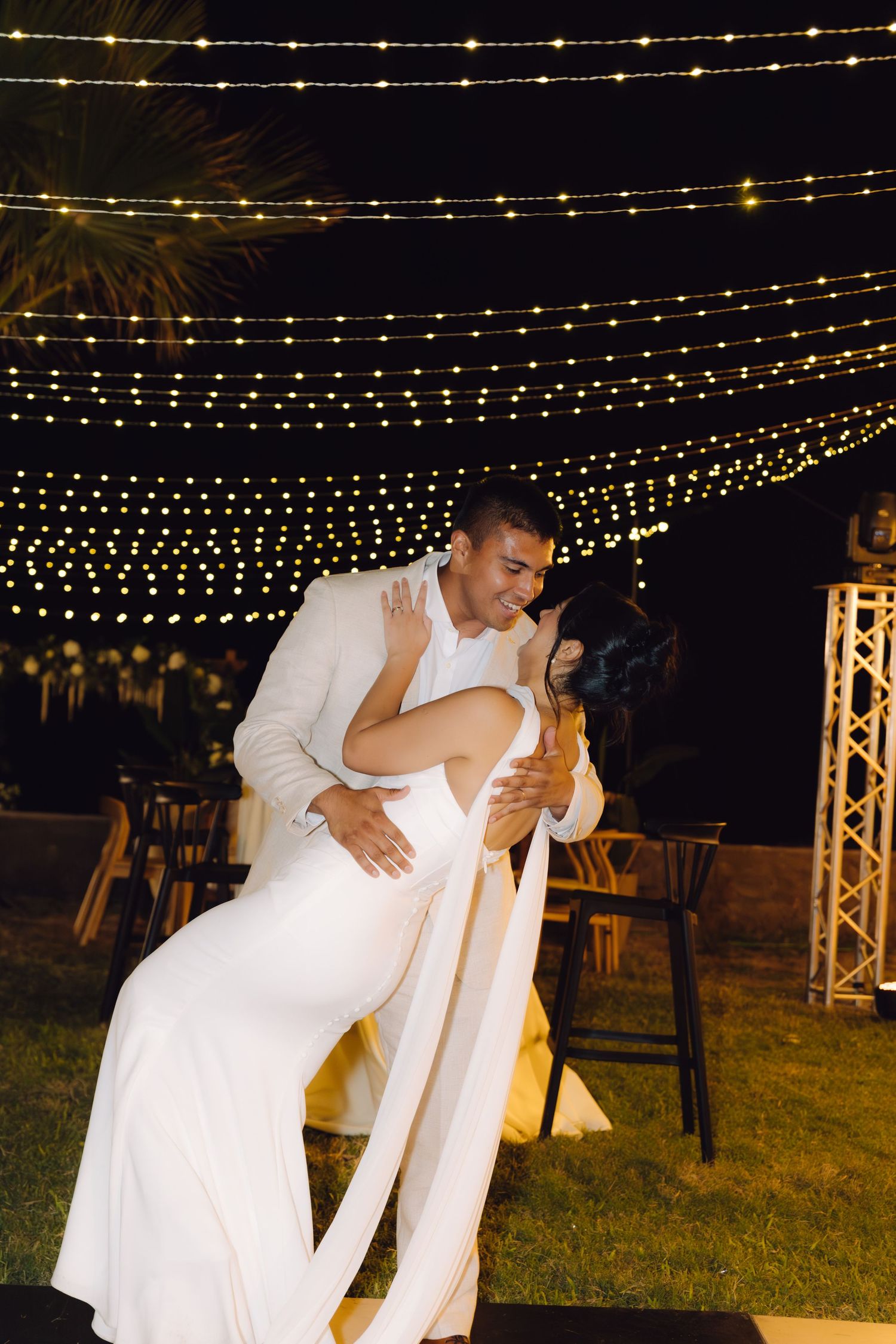 Groom dips bride in a white wedding gown under glowing string lights during their outdoor evening reception.