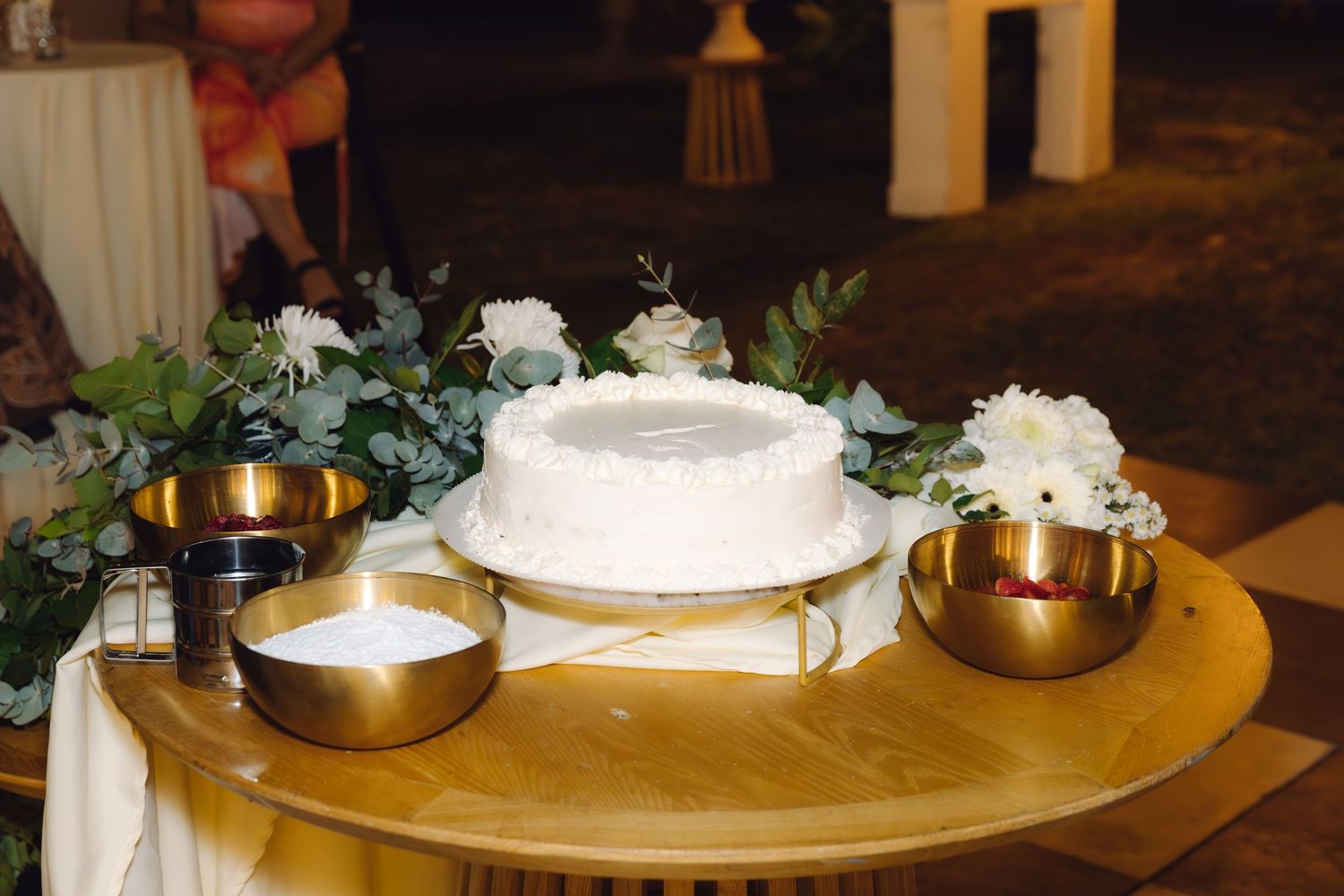 Elegant white wedding cake displayed on a wooden table with gold bowls and white floral arrangements.