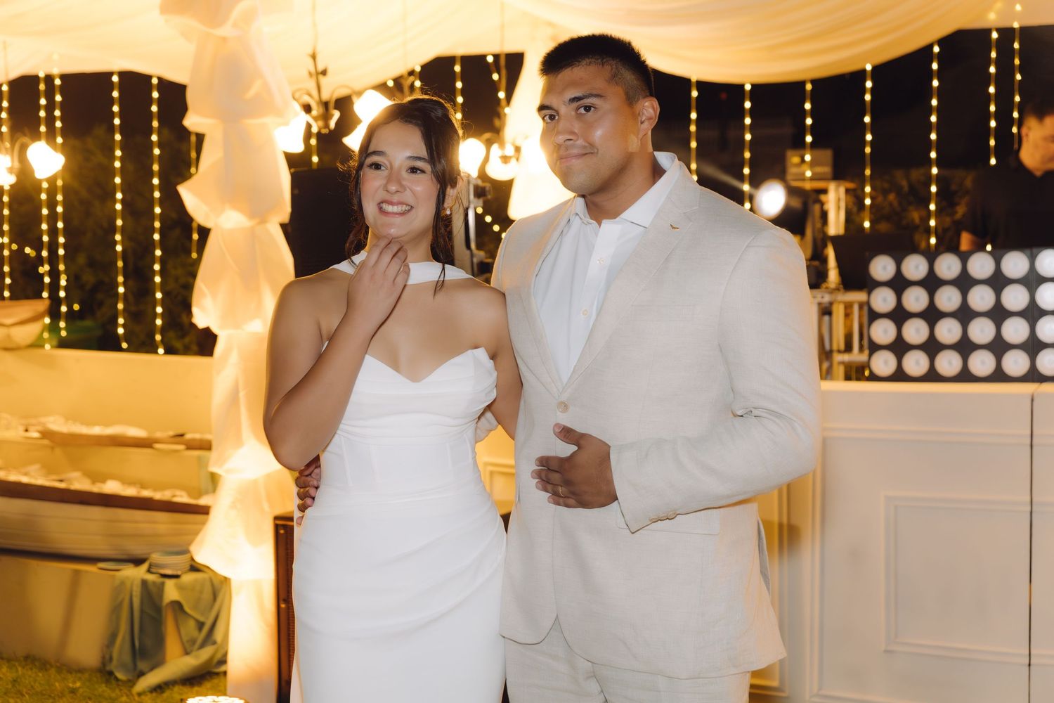 Bride in white strapless gown and groom in beige suit pose together at their outdoor wedding reception.