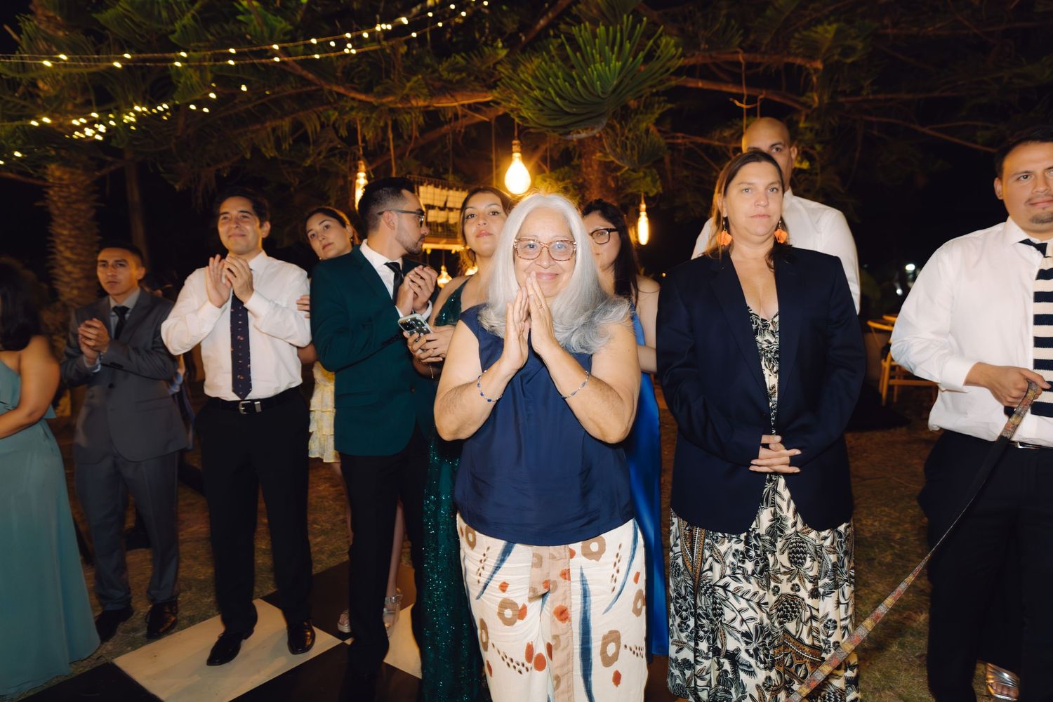 Wedding guests watch and applaud during an outdoor evening reception surrounded by tropical palm trees.
