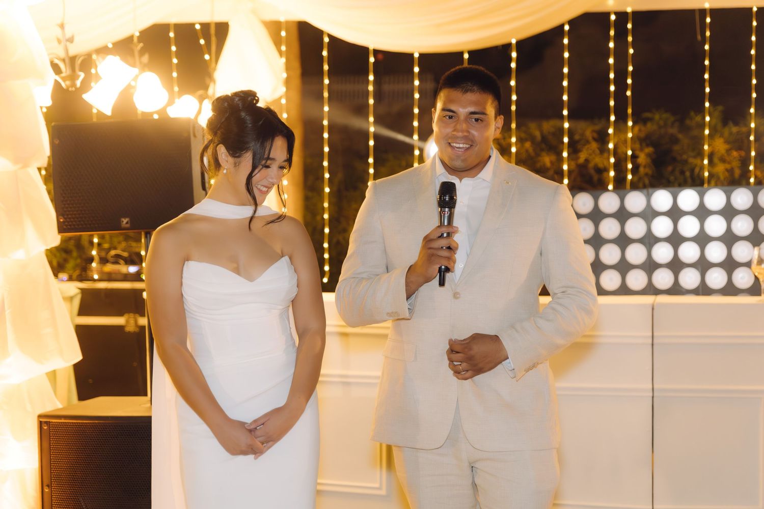 Groom in beige suit speaks into microphone beside bride in white gown at their outdoor wedding reception.