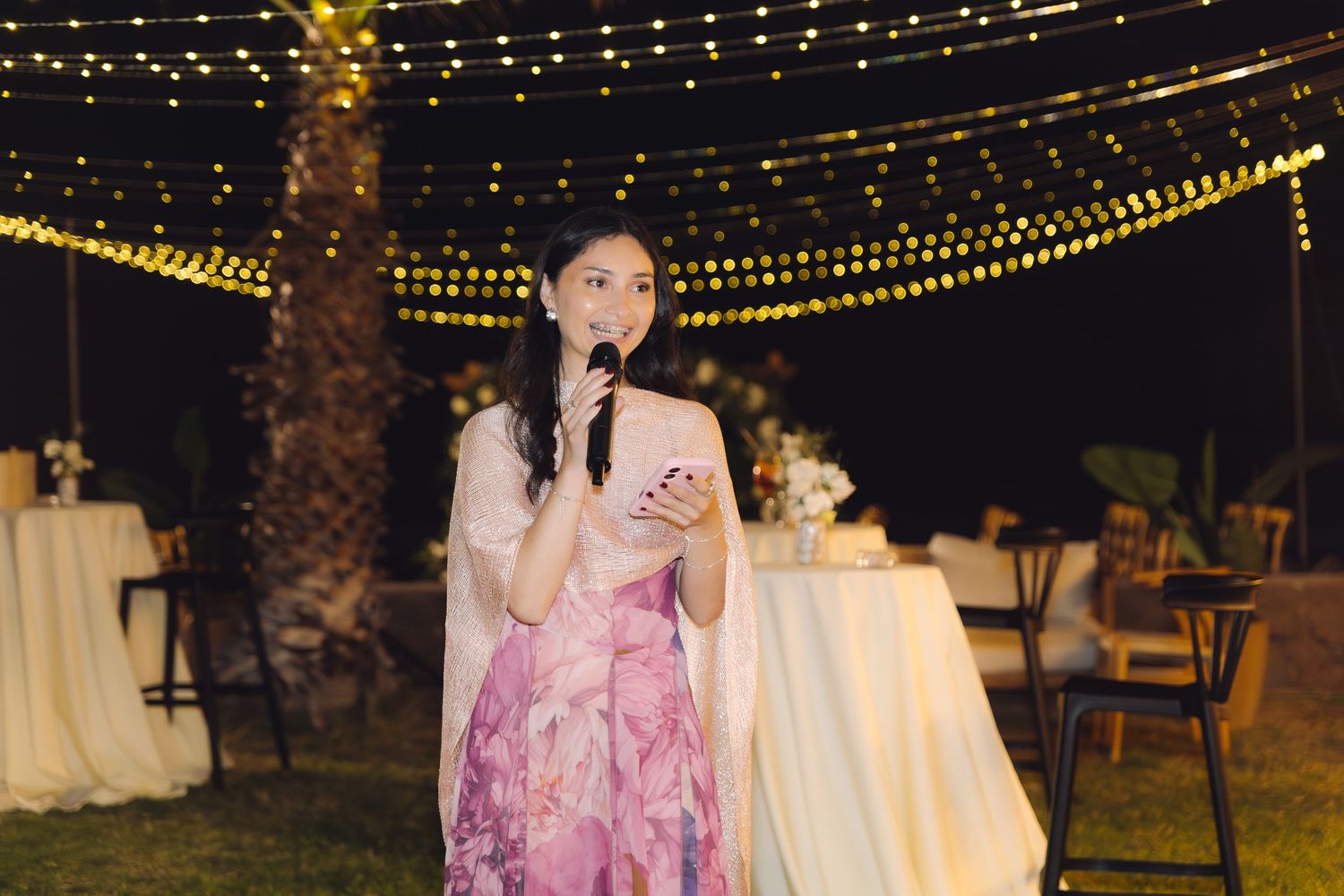Woman in pink floral dress delivers a heartfelt speech with microphone at an outdoor wedding reception.