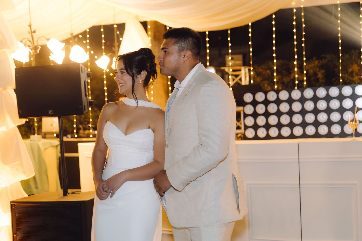 Bride and groom stand together smiling at their outdoor wedding reception with decorative string lights.