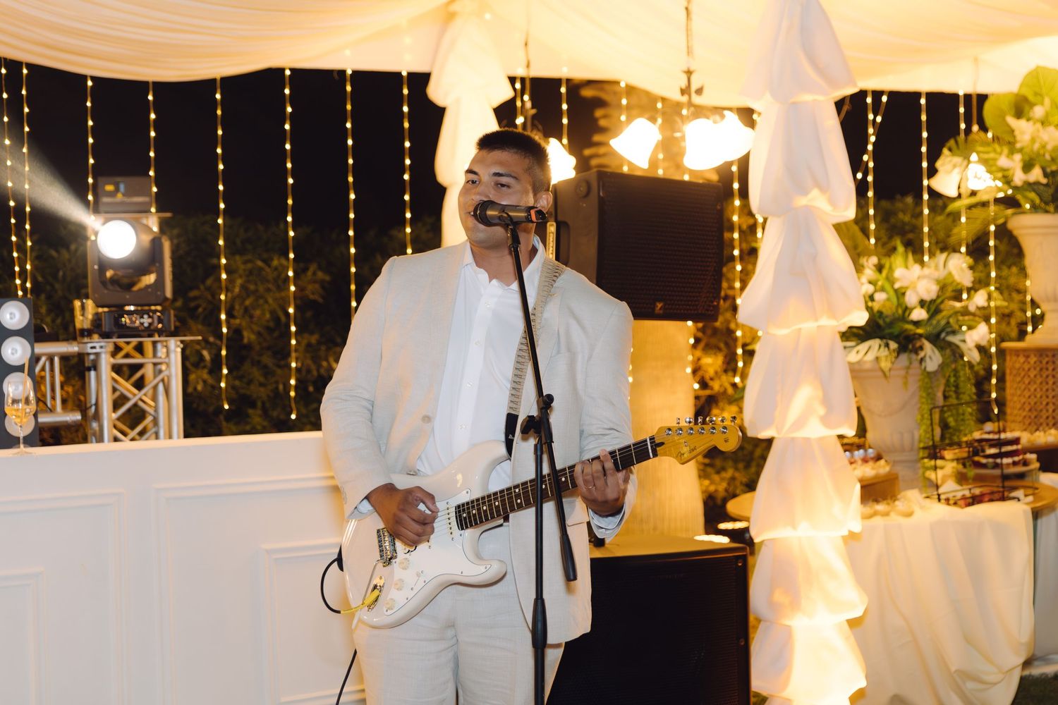 Man in white suit performs live music on electric guitar at an elegant wedding reception with fairy lights.