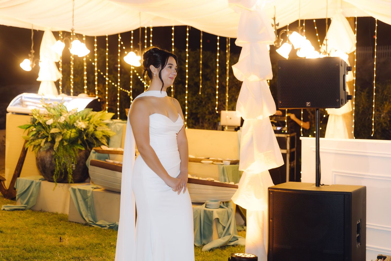 Bride in white strapless gown stands gracefully at outdoor wedding reception decorated with warm glowing lights.