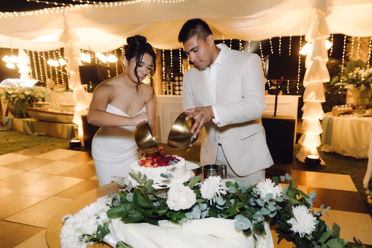 Smiling couple cuts berry-topped white wedding cake together at romantic outdoor wedding reception at night.