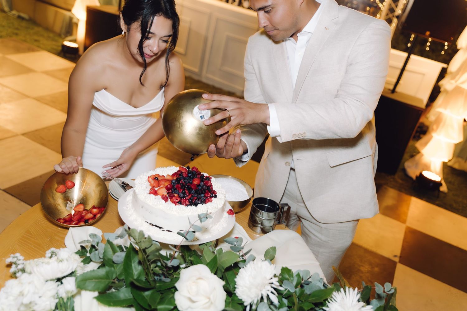 Bride and groom use golden bowl to top white berry wedding cake at evening reception.