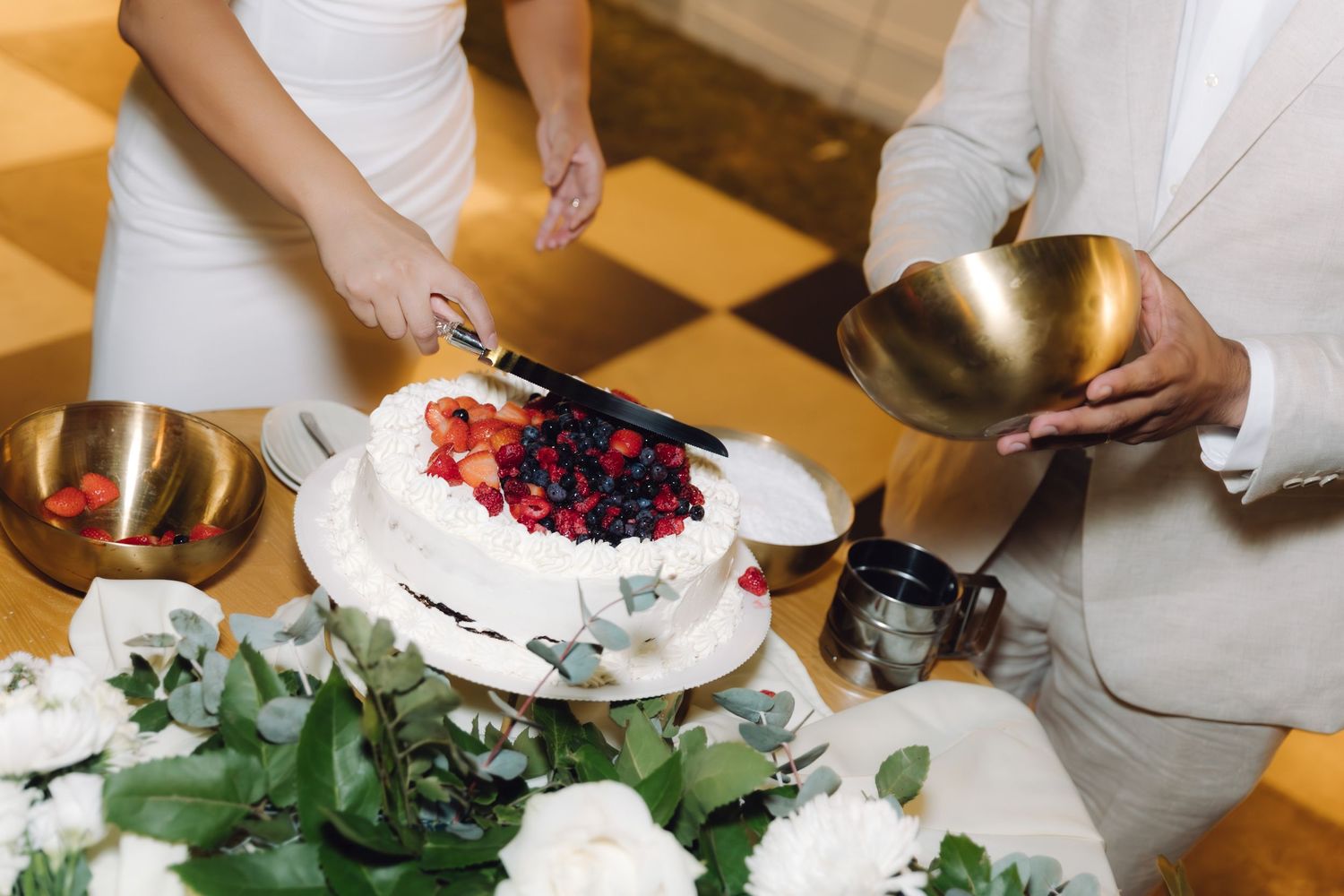 Close-up of couple cutting white wedding cake adorned with fresh strawberries and blueberries at reception.