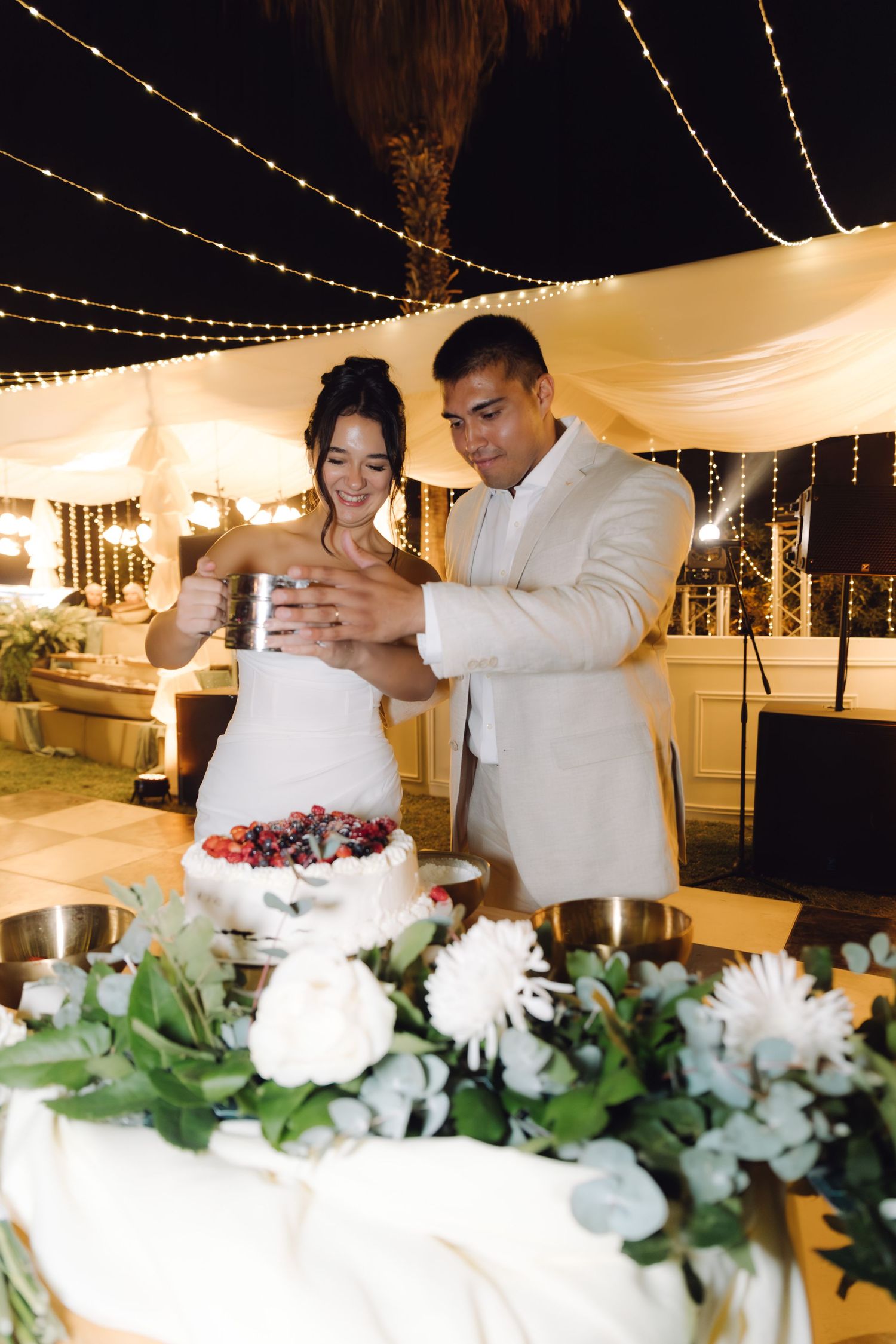 Laughing bride and groom dust wedding cake with powdered sugar at romantic outdoor evening reception.
