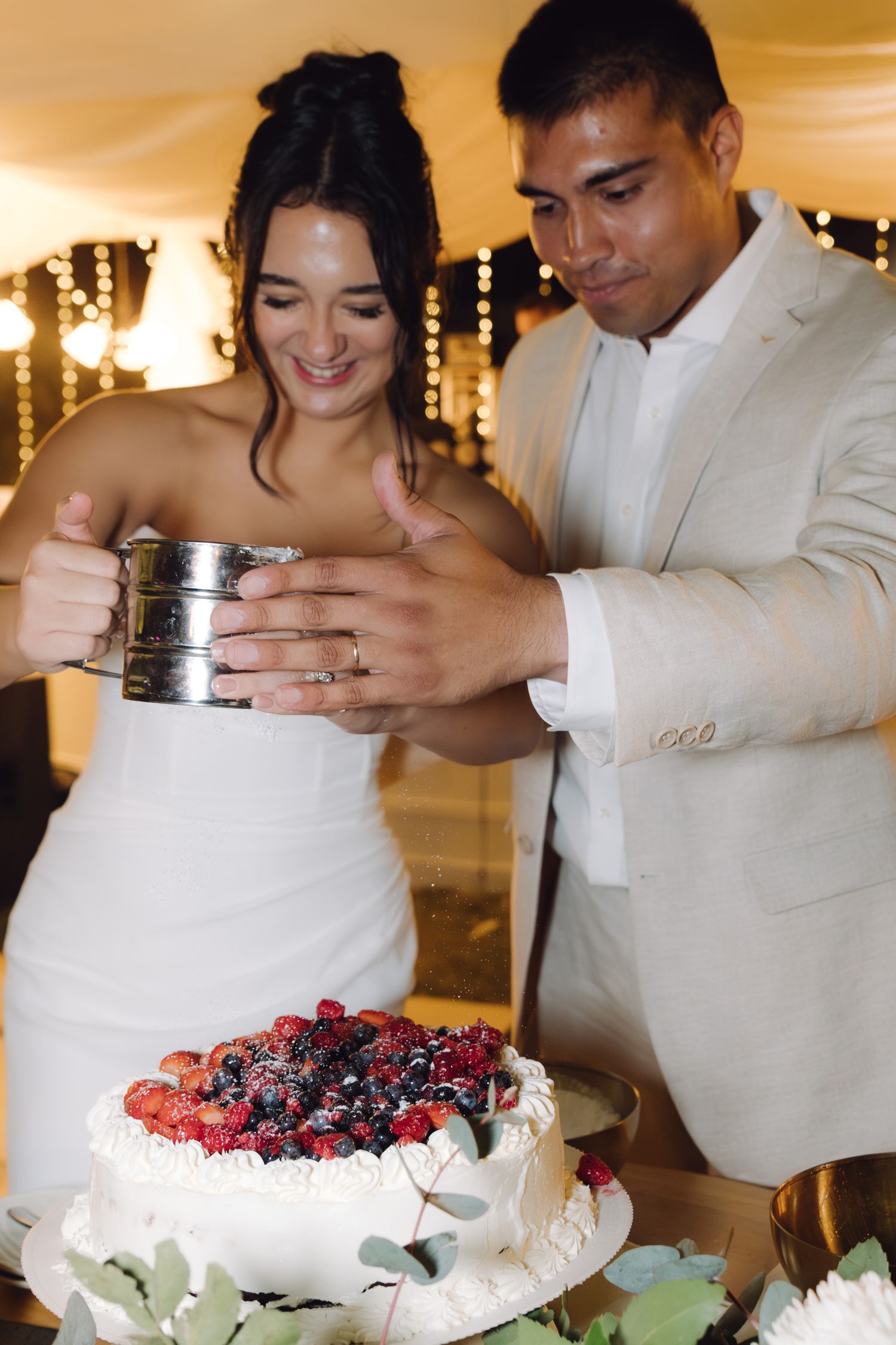 Bride and groom together dust berry-covered wedding cake with powdered sugar at elegant evening reception.