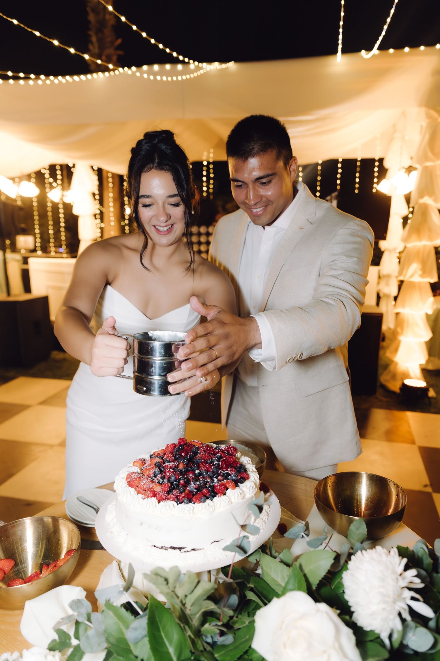 Couple smiles while sifting powdered sugar over berry-topped white wedding cake at night reception.