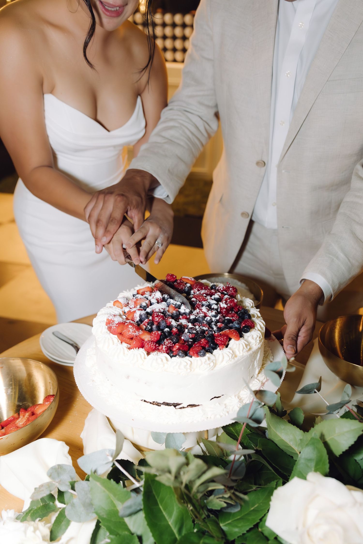 Bride and groom cutting a white wedding cake topped with fresh mixed berries and powdered sugar.