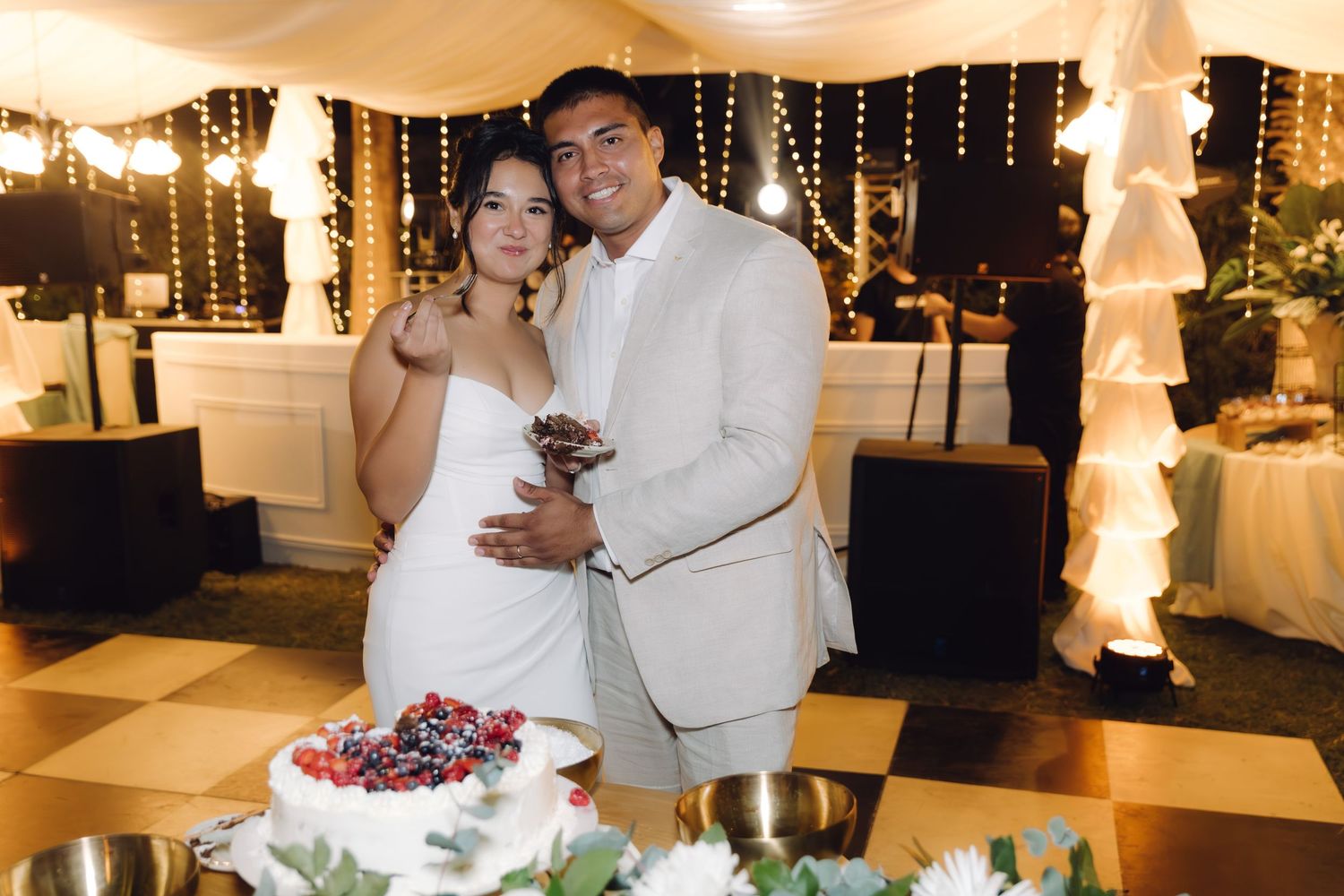 Smiling bride and groom posing together in front of their berry-topped white wedding cake at reception.