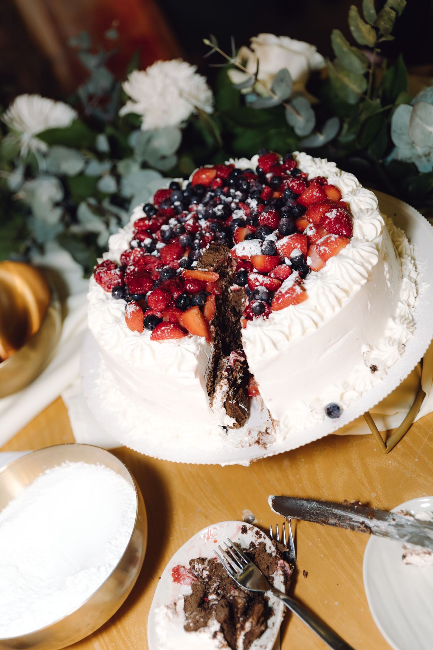 White wedding cake with mixed berry topping and a slice removed, displayed on a wooden table.