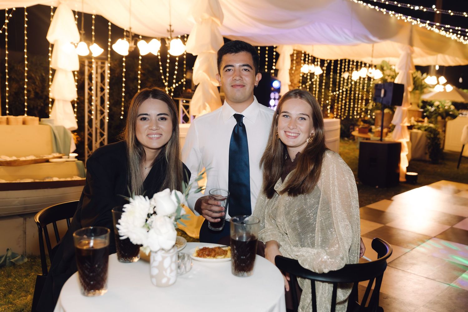 Two young women and a man in a tie smiling together at a decorated wedding reception table.