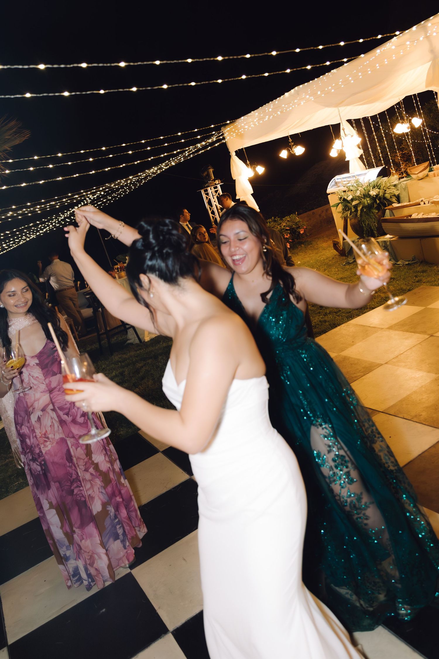 Bride in white gown dancing with a guest in an emerald sequin dress at an outdoor wedding reception.