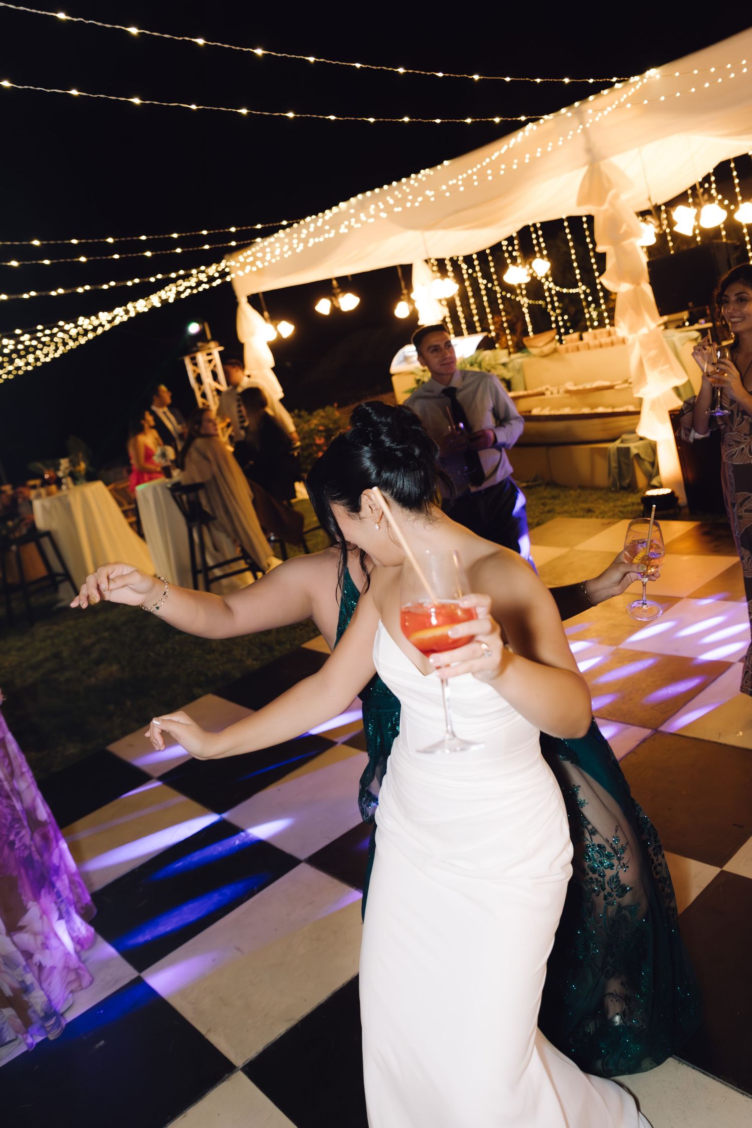 Bride and guest in emerald dress dancing on a black and white checkered floor at outdoor wedding.