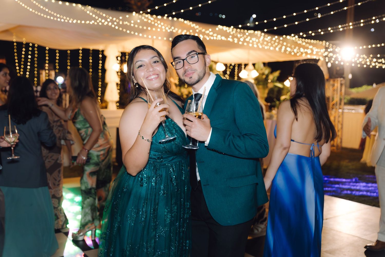Couple in formal attire raising champagne glasses and smiling at an outdoor evening wedding reception.