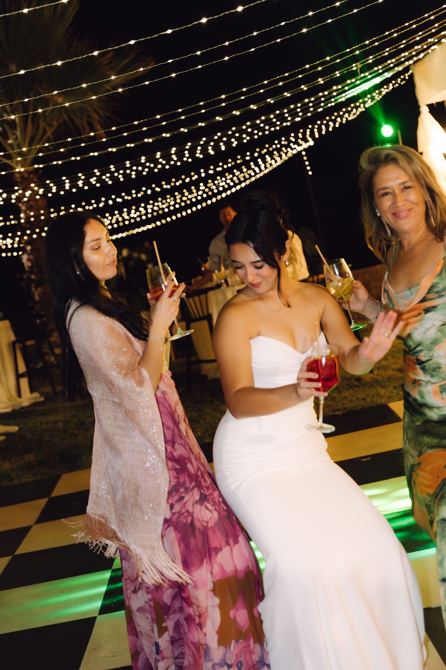 Bride in white strapless gown dancing and celebrating with two female guests at outdoor wedding reception.