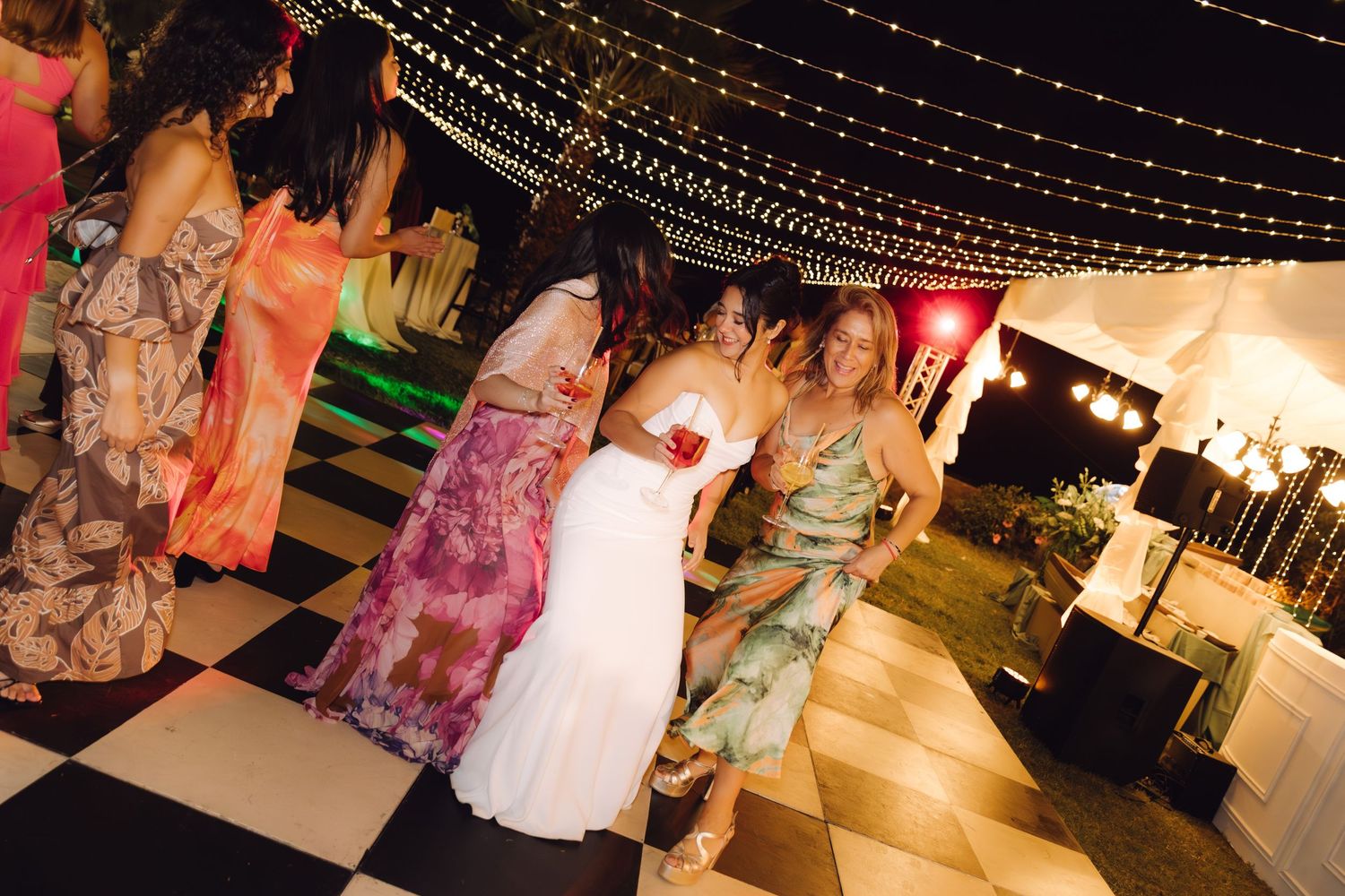 Bride in white gown dancing with guests on a checkered dance floor at an outdoor wedding reception.