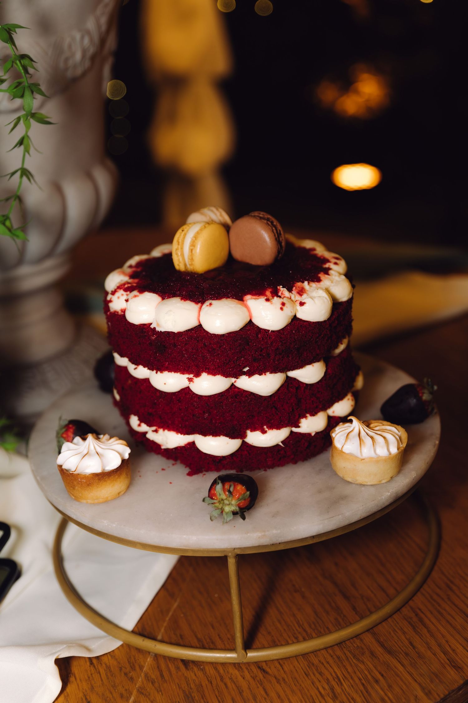 Elegant red velvet naked cake with white cream layers, macarons on top, surrounded by mini desserts on a marble stand.