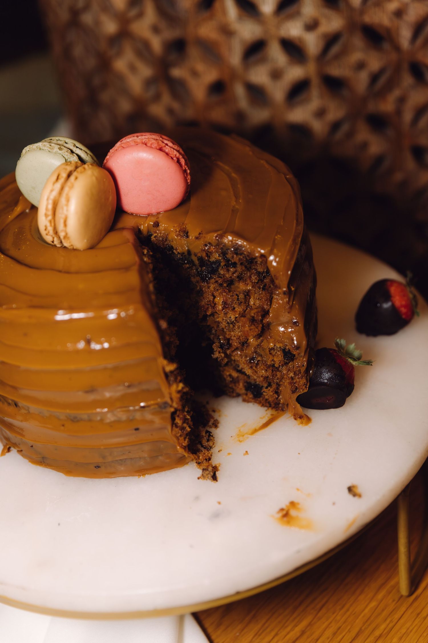 Sliced chocolate cake with caramel frosting topped with colorful macarons and chocolate-covered strawberries on white plate.