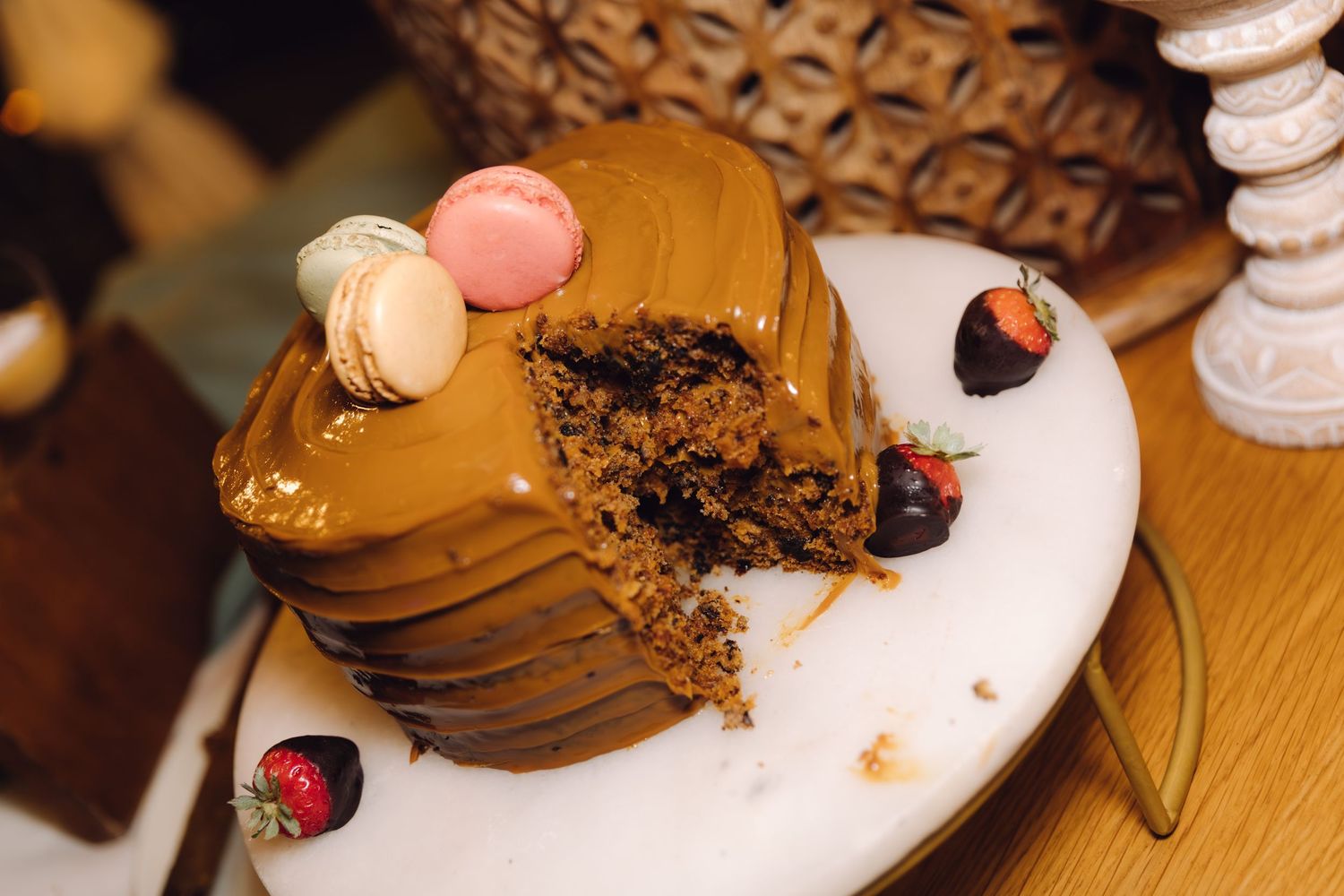 Close-up of partially eaten chocolate caramel cake with macarons and chocolate-covered strawberries on a white cake stand.