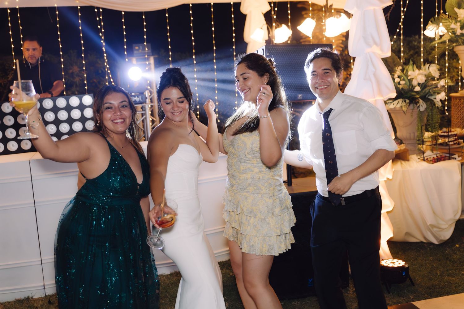 Four wedding guests smiling and celebrating on a festive outdoor dance floor with string lights at night.
