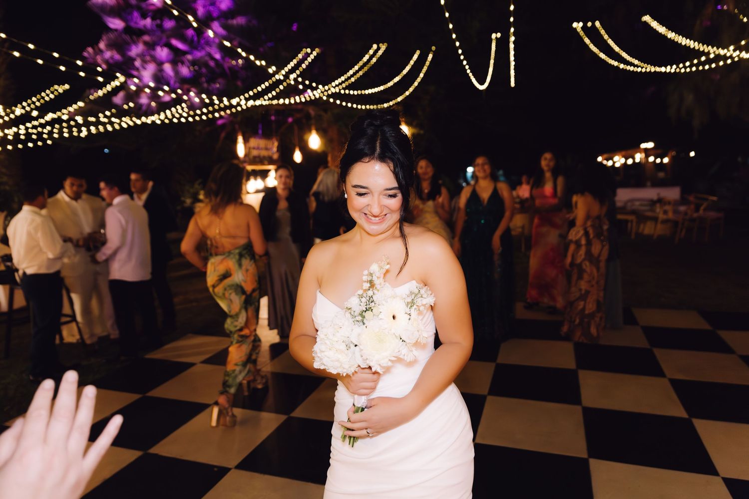 Radiant bride in strapless white gown smiles while holding white bouquet on black and white checkered dance floor at night.