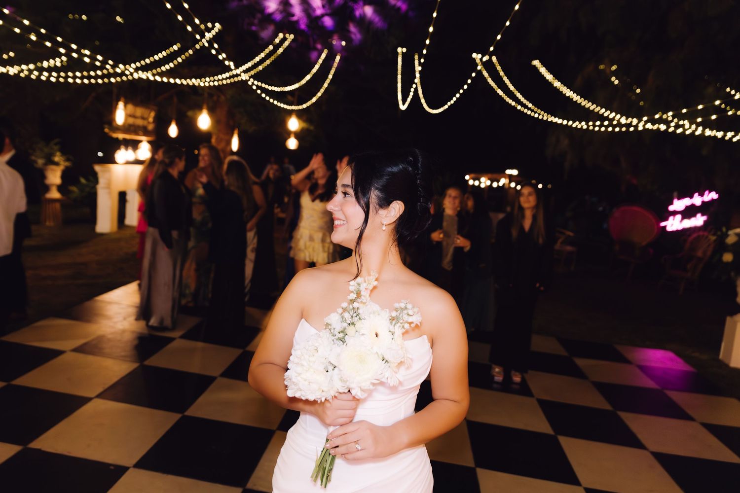 Bride in white gown holding bouquet standing on black and white checkered dance floor under string lights at night.