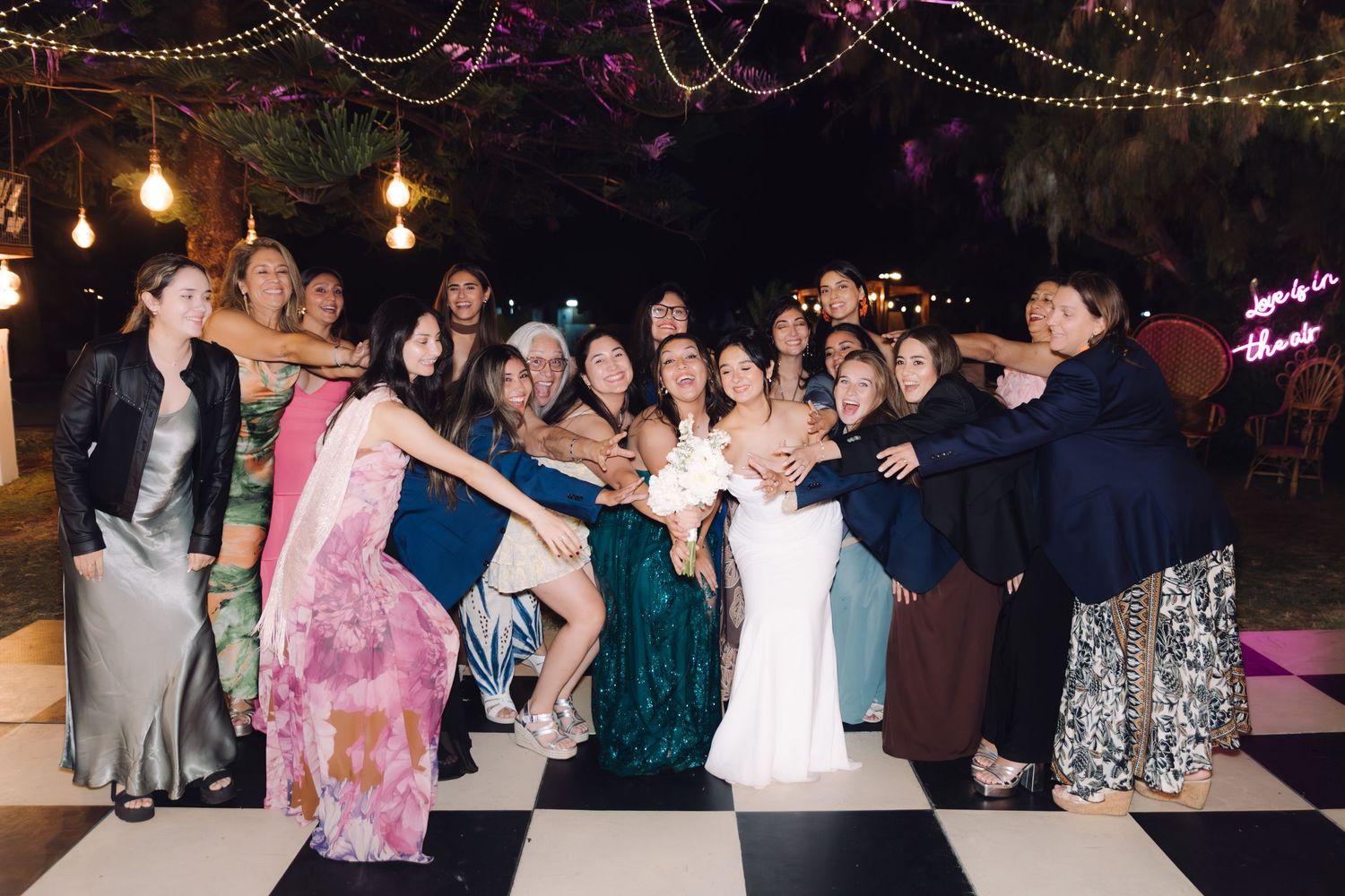 Bride surrounded by large group of celebrating female guests on checkered dance floor under string lights at outdoor wedding reception.