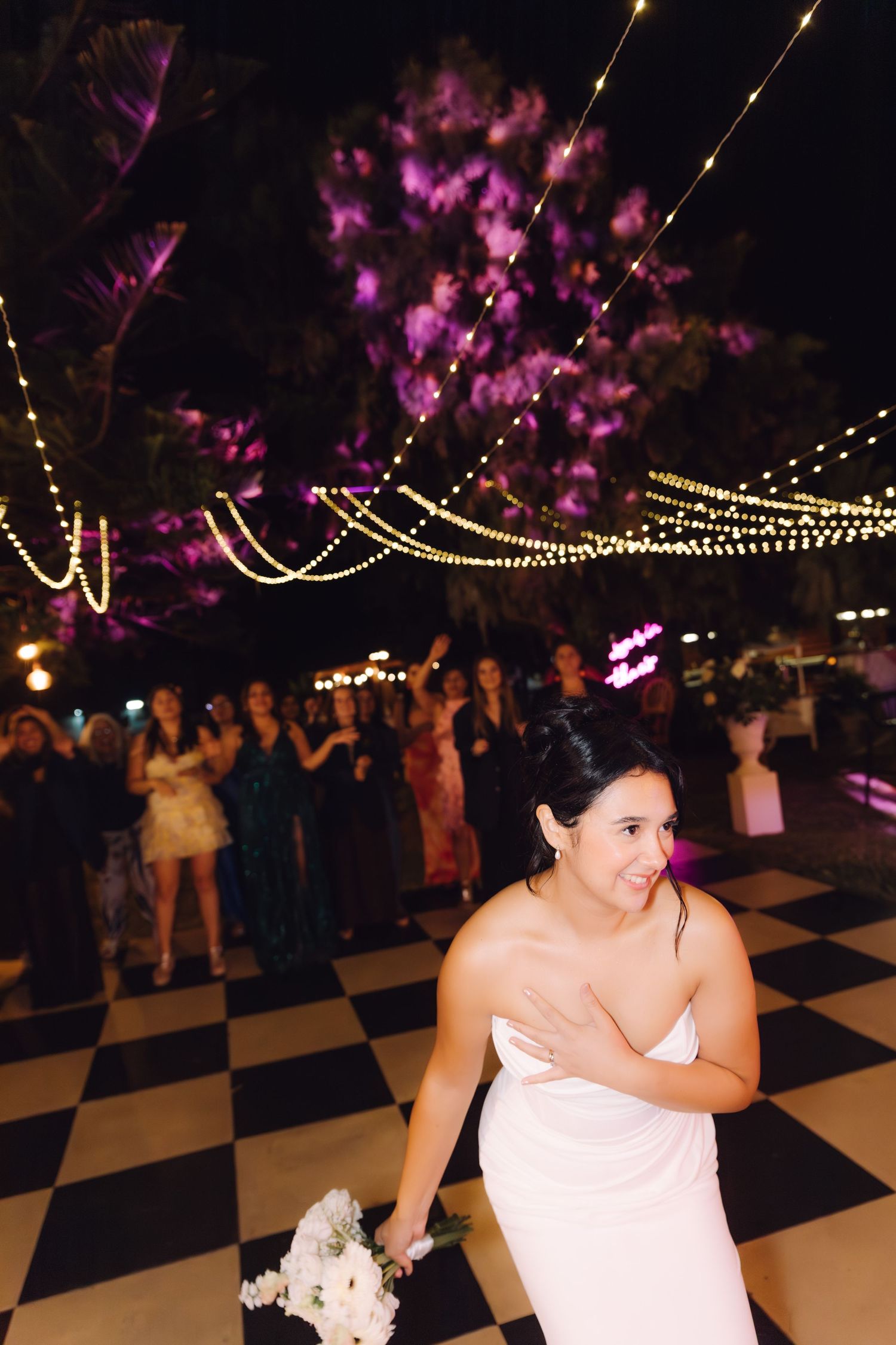 Bride preparing to toss bouquet of white flowers on checkered dance floor with guests watching under festive string lights at night.