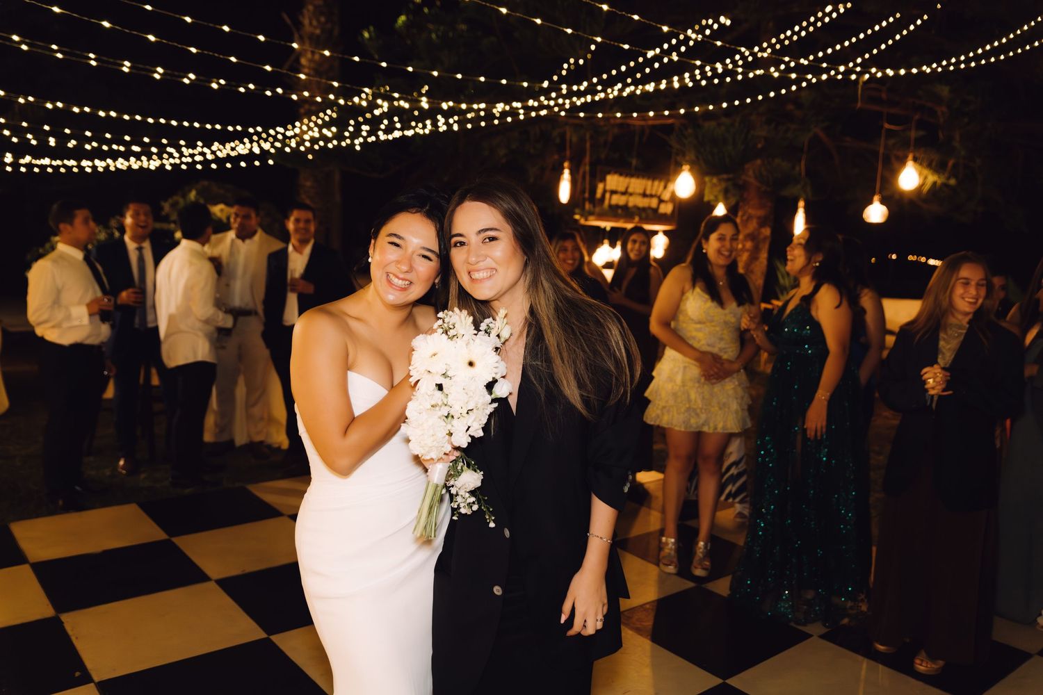 Bride in white gown holds bouquet posing with guest on checkered dance floor under string lights at outdoor wedding reception.