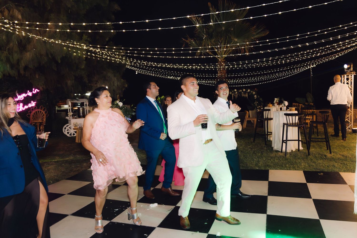 Guests dancing joyfully on checkered dance floor under string lights at elegant outdoor evening wedding reception.