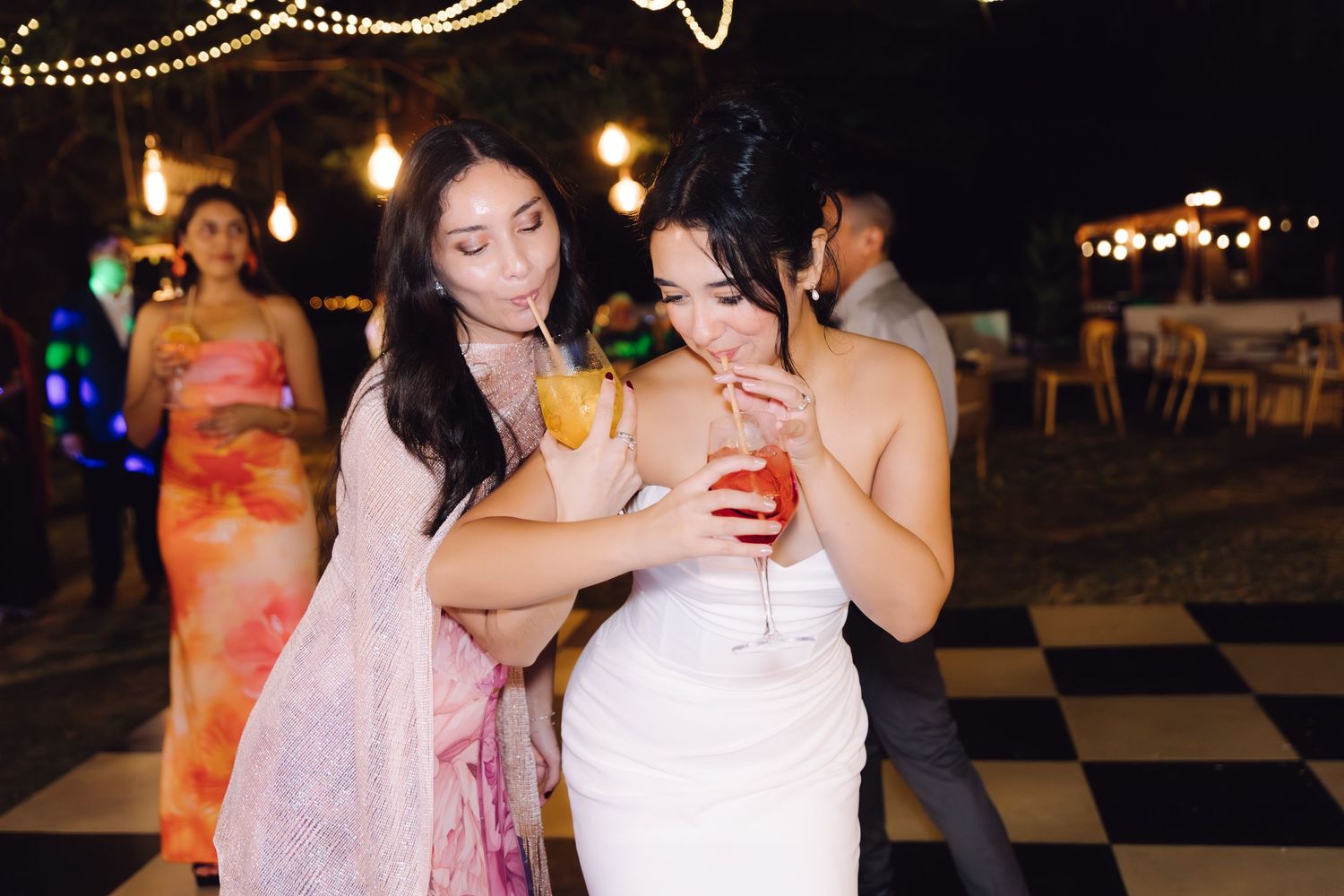 Bride in white dress shares cocktails with guest in pink dress on checkered dance floor at outdoor wedding.